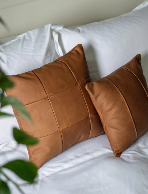 A Large Leather Pillow by Urban Southern rests on a neatly made bed with white sheets. In the foreground, a green plant is visible, slightly out of focus.