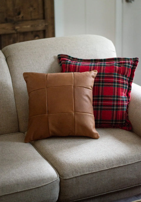 A beige sofa holds two accent pillows: the Urban Southern Large Leather Pillow with a grid pattern in front and a red plaid cushion behind. A wooden cabinet and white wall are in the background.