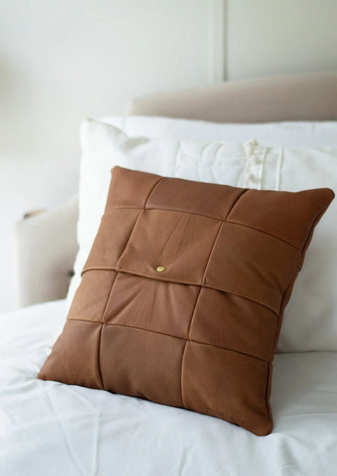 The Urban Southern Large Leather Pillow, featuring a patchwork grid design and central button detail, adds a striking accent to a white bed with its bold look against the light headboard.