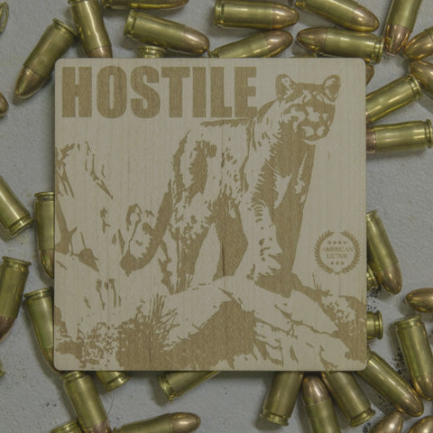An American Lictor Wood Sticker featuring a square wooden design with an engraved mountain lion and the word HOSTILE sits on a gray surface surrounded by scattered bullets.