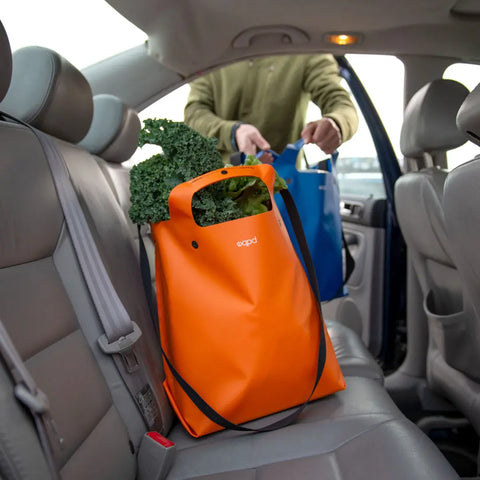 A person places grocery bags, including a durable eqpd LastBag - Shoulder in bright orange filled with leafy greens, onto the back seat of a car with gray leather interior.