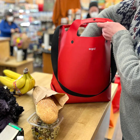 A shopper places bananas, a loaf of bread, and salad into an eqpd LastBag - Shoulder made in Twisp, setting it on the counter with other customers in the background.