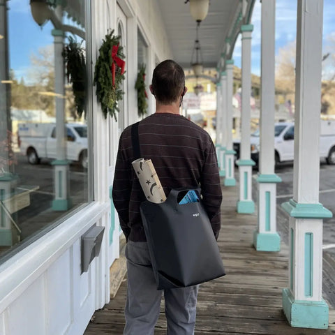 A person with short hair in a striped sweater stands on a wooden sidewalk, carrying an eqpd LastBag - Shoulder and a star-patterned mat. Wreaths and shop windows line the charming Twisp street.
