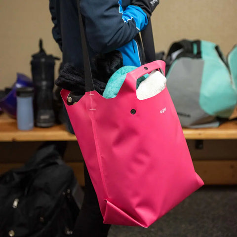 In a locker room, someone carries the eqpd LastBag - Shoulder in bright pink, packed with clothes and a towel. On a wooden bench behind them are more bags, gym gear, and a water bottle. This durable bag is ready for any routine.
