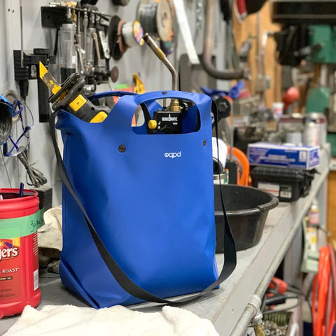 The eqpd LastBag - Shoulder, a durable blue everyday bag made in Twisp, rests on a busy workshop bench amid hardware, equipment, and containers.
