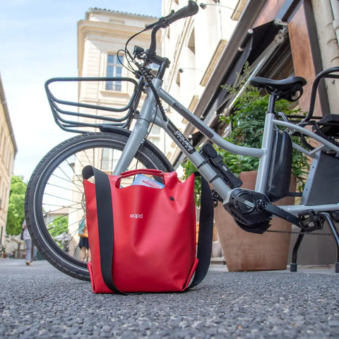 A grey bicycle is parked on a city street, with an eqpd LastBag - Shoulder in bright red and black straps standing upright on the ground in front of it. Buildings and plants are visible in the background.