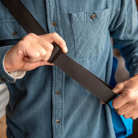 A person in a blue button-up adjusts the black strap of their eqpd LastBag - Shoulder, a durable everyday bag made in Twisp, worn across their shoulder.