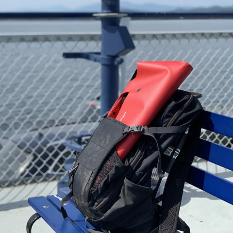 An eqpd LastBag - Shoulder, black and durable, with a red kickboard peeking out, rests on a blue bench by a chain-link fence overlooking calm water on a sunny day.