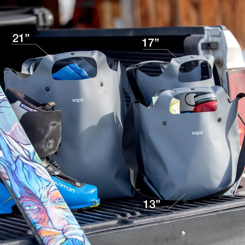 Three eqpd LastBag - Shoulder bags in gray (sizes 21, 17, and 13), made to last in Twisp, sit in a truck bed with ski gear—colorful skis and a blue boot—showcasing their waterproof durability.