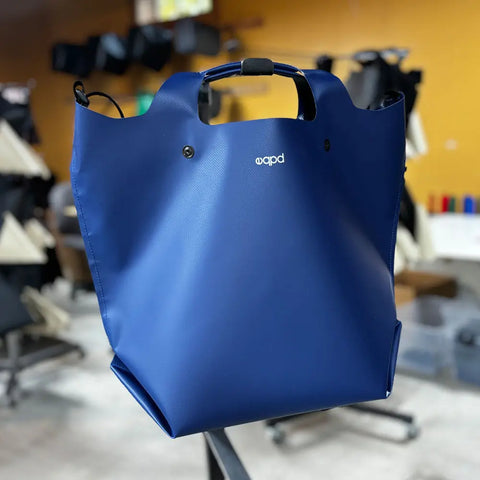 The eqpd LastBag - Shoulder is a large blue structured tote with short handles and a white apd logo, shown on a metal surface in a workshop. Its modern minimalist design and durable build make it the perfect bag for any adventure.