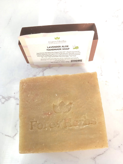 A square bar of natural soap embossed with Forev'Herbs sits on a white surface. Behind it, the Lavender Aloe Handmade Soap packaging lists ingredients, ideal for sensitive skin.