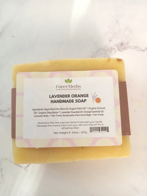 A rectangular bar of Forev'Herbs Lavender Orange Handmade Soap with a white and lavender label listing ingredients and directions. This yellow lavender essential oil soap is perfect for women seeking a natural, handmade touch.