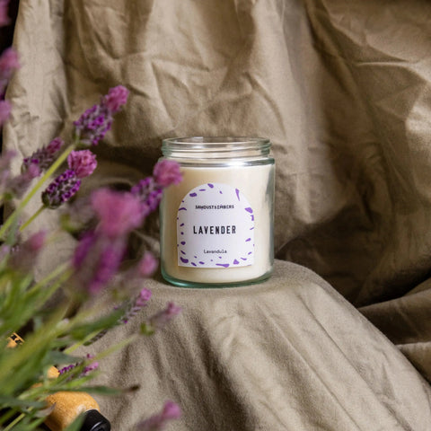 The Sawdust & Embers Lavender - Signature Candle sits on beige fabric with lavender flowers, offering an inviting touch of premium home fragrance to any space.