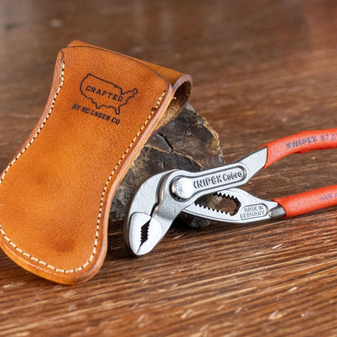 A KC Laser Co USA-made leather belt holster for Knipex 5 pliers is shown on a wooden surface next to red-handled pliers. The handmade pouch is crafted from veg-tanned cowhide and stamped with an outline of the USA.