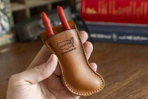 A hand holds the Leather Belt Holster for Knipex 5 Pliers by KC Laser Co, a handmade, veg-tanned cowhide tool pouch engraved with the brand name. Blurred books and objects are visible in the background.