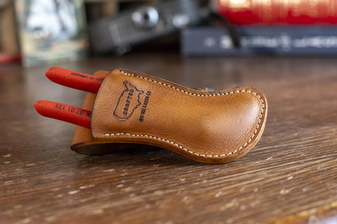 A handmade Leather Belt Holster for Knipex 5 Pliers by KC Laser Co holds two red-handled tools on wood. Stamped “CRAFTS” and “STEEL BLADE,” this USA-made pouch sits before blurred books and objects in the background.