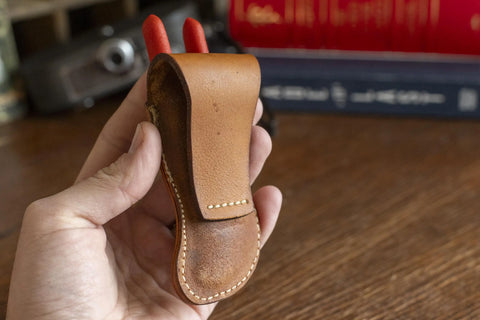 A hand holds the KC Laser Co Leather Belt Holster for Knipex 5 Pliers, a handmade veg-tanned cowhide tool pouch with visible stitching. Made in the USA, this stylish sheath rests on a wooden surface with blurred books in the background.