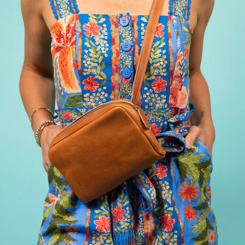 Wearing a colorful floral dress, a person holds the CASUPO Leather Fanny Pack - Chocolate, crafted from sustainable brown leather, while standing against a light blue background.