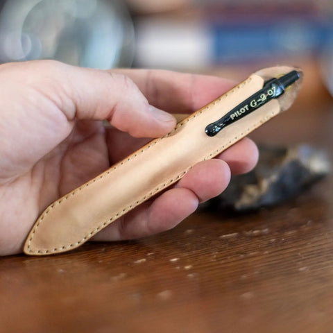 A hand holds a KC Laser Co Leather Pen Sleeve for G2 Pens, handcrafted in the USA, with a black Pilot G-2 07 pen clipped to it and resting on wood—ideal for those valuing style and quality.