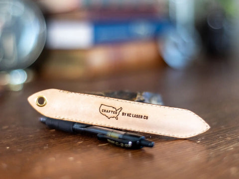 A handcrafted tan leather pen sleeve for G2 pens by KC Laser Co, stamped with CRAFTED BY KC LASER CO and a USA outline, displays quality American craftsmanship—shown here beside a complimentary G2 pen.