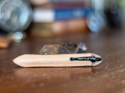 A black Pilot G2 pen in a KC Laser Co Leather Pen Sleeve – handcrafted in the USA by Crafted x KC Laser Co – lies on a wooden surface, blurred books and objects in the background.