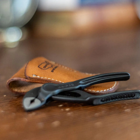A pair of black Knipex 4 XS pliers rests on a wooden surface beside the KC Laser Co Leather Tool Holster—a tan, veg-tanned cowhide belt pouch—while the blurred background highlights these USA-made tools in the foreground.
