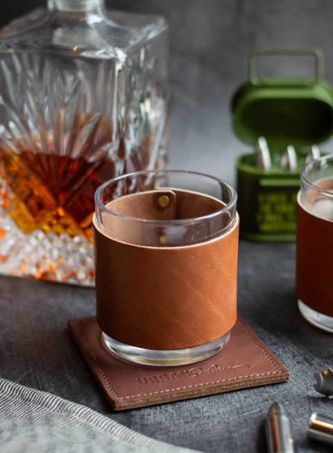 The Urban Southern Leather Wrapped Whiskey Glass sits on a matching coaster next to a crystal decanter and green case with metal accents—an elegant addition to any bar cart.