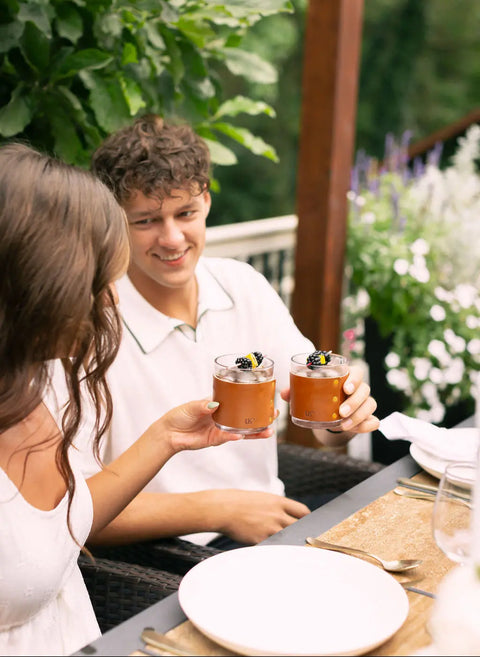 Two people sit outdoors, smiling and holding up Urban Southern Leather Wrapped Whiskey Glasses with brown cocktails garnished with blackberries—a perfect bright, summery scene for relaxing by a stylish bar cart surrounded by lush plants and flowers.