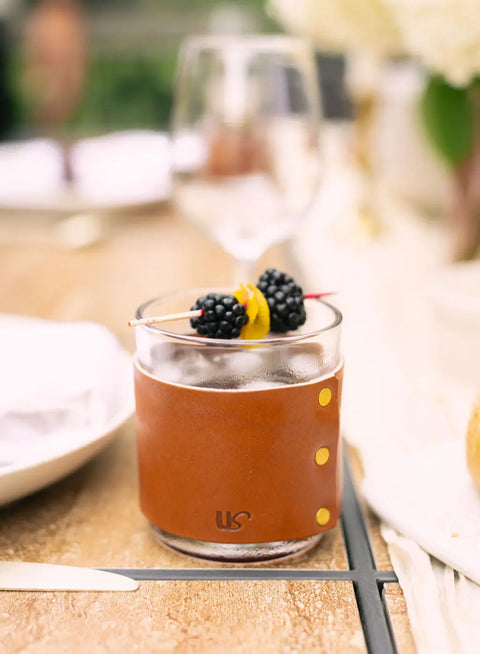 Enjoy a cocktail served in the Urban Southern Leather Wrapped Whiskey Glass, garnished with two blackberries and a lemon twist, set on a table with softly blurred dining items in the background.