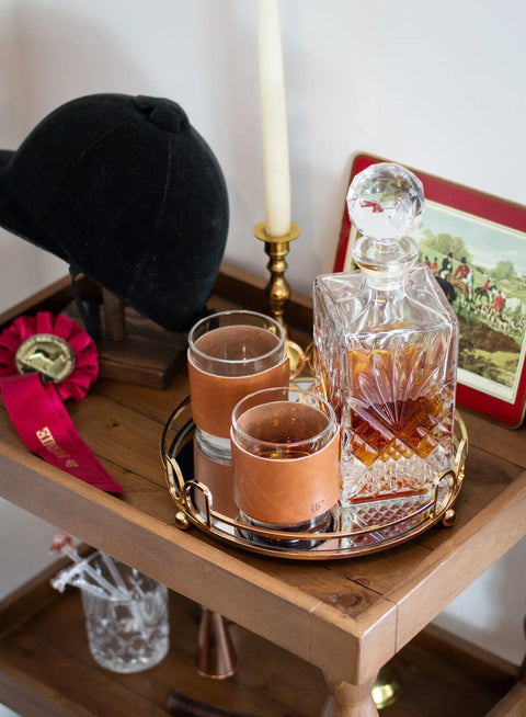 A wooden bar cart displays a glass decanter, two Urban Southern Leather Wrapped Whiskey Glasses, a black equestrian helmet, red ribbon, candle, and a framed horse-riding photo.
