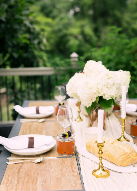 An outdoor dining table set features white plates, gold utensils, wine glasses, a Leather Wrapped Whiskey Glass by Urban Southern, white napkins with brown rings, bread loaf, white flowers in a vase, gold candle holders, dessert cups, and greenery.