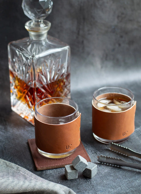 An Urban Southern Leather Wrapped Whiskey Glass sits beside a crystal decanter of whiskey, one glass with ice. Gray whiskey stones, metal tongs, and a leather coaster are arranged nearby on a gray bar cart.