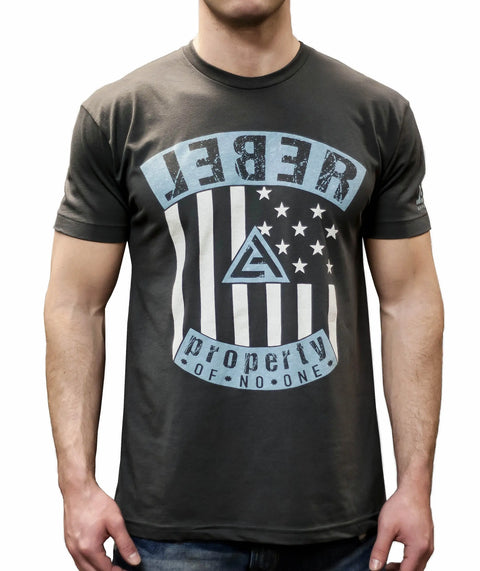 A man wearing The Commander t-shirt from Leber Apparel Line, featuring a distressed REBEL graphic with an abstract triangle, stars, stripes, and property of no one—ideal for fans of patriotic style.