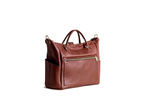 The Urban Southern Legacy Backpack Tote, a brown leather bag with gold hardware, two top handles, convertible straps, a front zip pocket, and side pockets, is shown in heritage style against a white background.