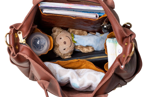An open Urban Southern Legacy Backpack Tote in brown leather, viewed from above, holds a stuffed monkey toy, baby bottle, diapers, folded blanket, papers or magazines, and a black zipped pouch with convertible straps.