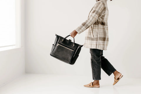 A person in a plaid coat, black jeans, and tan flats walks indoors with Urban Southern’s Legacy Backpack Tote. Soft natural light on a white background emphasizes the brand’s timeless heritage style and the bag’s versatile, convertible straps.