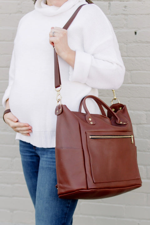 A pregnant woman in a white sweater and jeans stands before a light brick wall, carrying Urban Southerns Legacy Backpack Tote featuring gold hardware and convertible straps, showcasing the brand’s heritage style.