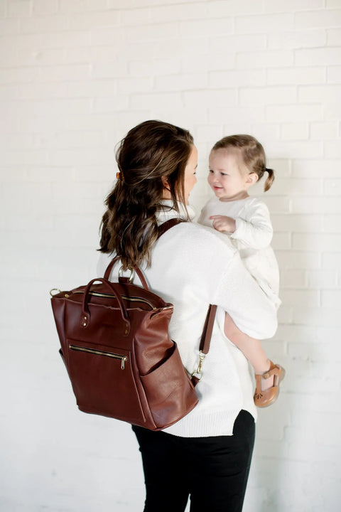 A woman in a white sweater holds a smiling toddler against a white brick wall, carrying the Urban Southern Legacy Backpack Tote with convertible straps, reflecting the brand’s heritage style.