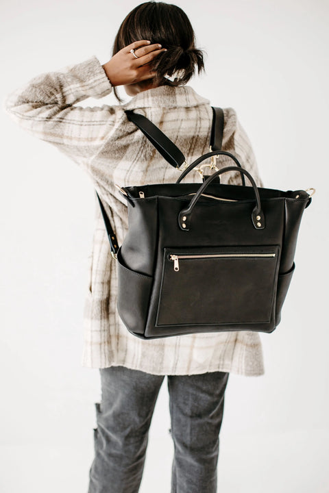 Wearing a plaid coat and gray jeans, a person stands facing away, carrying the Urban Southern Legacy Backpack Tote—a large black leather bag with convertible straps and a front zip pocket—resting one hand on their head against a white background.
