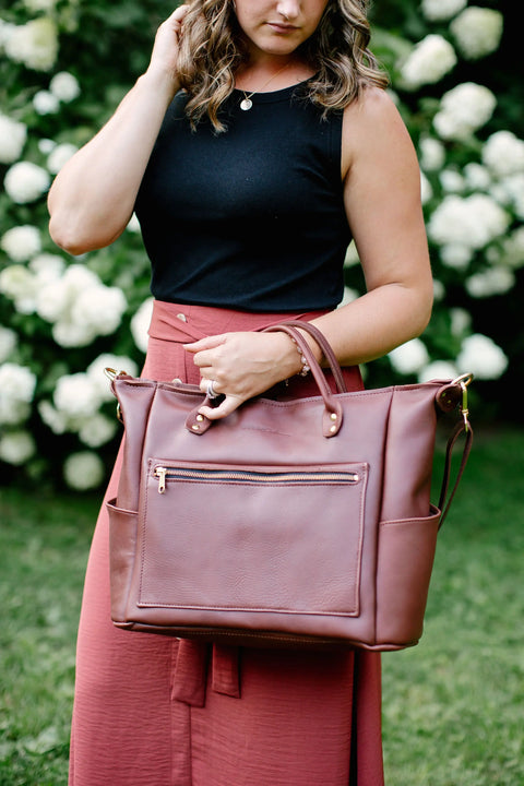 A woman in a sleeveless black top and rust skirt stands outdoors holding Urban Southern’s Legacy Backpack Tote with convertible straps. Blurred white flowers and greenery appear in the background.