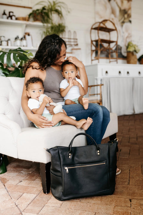 A woman sits on a white couch with two young children in her lap. The Urban Southern Legacy Backpack Tote with convertible straps sits on the floor, bringing heritage style to the cozy living room.