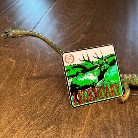 A toy lizard on a wooden floor displays the American Lictor Sticker: Elk - Legendary—a vibrant 3x3 sticker featuring an elk and bold red LEGENDARY text, perfect for celebrating this majestic AF moment.