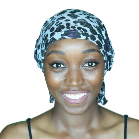 A woman smiles warmly, wearing the black and white leopard-print Chemo Beanies® - Katy headscarf by Chemo Beanies®, paired with a black strap top, against a plain background.