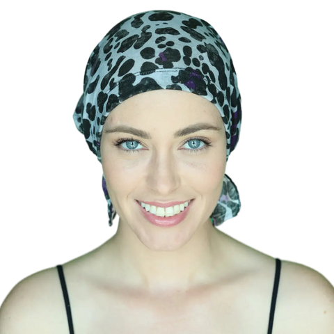 A woman with light skin and blue eyes smiles at the camera, wearing a black strap top and the Chemo Beanies® Katy scarf in a black and white pattern from Chemo Beanies®, set against a plain background.