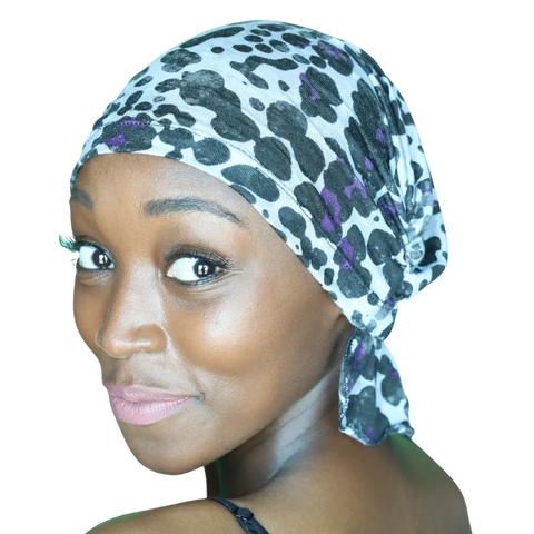 A woman with medium-dark skin smiles over her shoulder, wearing the Chemo Beanies® Katy—a black, white, and purple leopard-print scarf—showcasing how stylish chemo head covers from Chemo Beanies® can look against a plain backdrop.