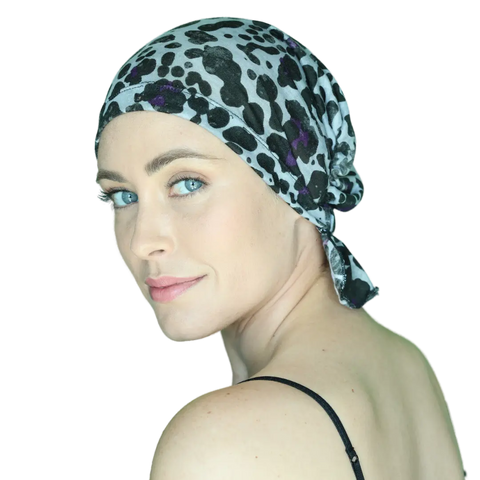 A woman with light skin and blue eyes models the Chemo Beanies® Katy chemo beanie scarf, glancing over her shoulder and smiling softly in a black strap top against a plain background.