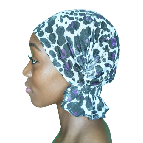 A woman in profile wears the Chemo Beanies® Katy scarf—a white chemo head cover with black and purple leopard print—elegantly tied at the back. She has smooth brown skin and long lashes, set against a plain black background.