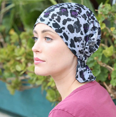 A woman in a pink top wears the Chemo Beanies® Scarves - Katy in black and white leopard print, sitting outdoors and gazing thoughtfully to the left with green plants behind her.