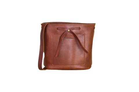 The Lexington Bow by Urban Southern is a brown leather shoulder bag featuring a front pocket, leather bow accent near the top, and long crossbody strap, shown against a plain white background.