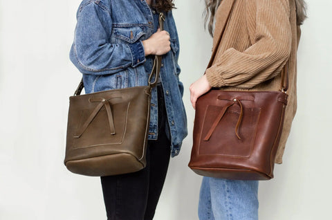 Two people stand side by side, each carrying The Lexington Bow crossbody from Urban Southern—one in brown and one in olive green. One wears a blue denim jacket, the other a tan corduroy jacket.